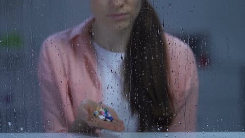 Lonely Sick Woman Holding Palm of Medication Near Rainy Window Looking to Camera