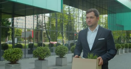 Businessman Gets Fired and Walking Near Office Center with Carton Box and Documents with a Table