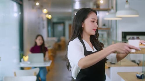 Die attraktive asiatische Caféangestellte trägt eine Schürze und gibt dem Kunden eine Lebensmitteltüte zum Mitnehmen.