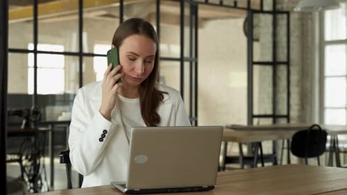 Woman Sits at a Desk in the Office and Talks on the Phone and Takes Notes at the Same Time