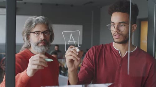 Deux hommes d'affaires d'âge mixte dessinant sur un mur de verre et parlant au bureau