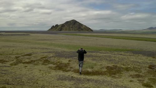 Aerial drone view tracking a person trekking over a flat, green field, towards a hill, in Iceland