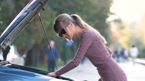 Young Upset Woman Driver Talking on Mobile Phone Near a Broken Car with Open Hood Waiting for Help