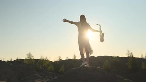 Slender Woman in White Dress Stands on Top of Mountain Hands to the Side Holding Saxophone in One