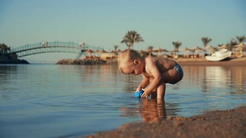 Funny Baby Boy Pouring Water Into Blue at Calm Sea Bay in Evening Time.
