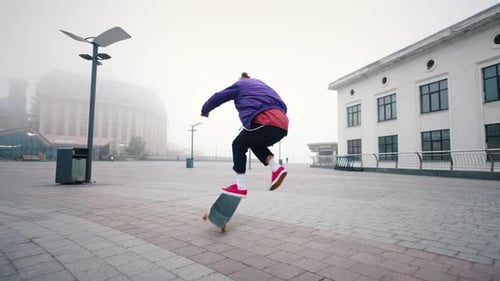 Rear View of Young Modern Guy Training His Skateboarding Skills in an Empty City Square on a Foggy
