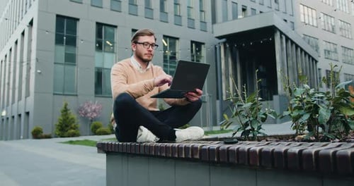 Joven usando laptop sentado al aire libre cerca del fondo del edificio de la empresa de oficinas.
