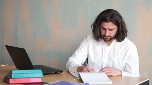 Young Beraded Man with Long Hair in White Shirt Working at Office with Papers and Laptop
