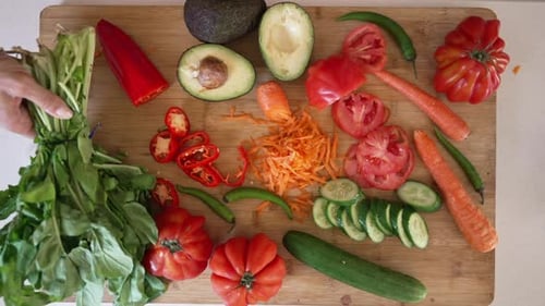 Top View Wooden Cutting Board with Tomato Cucumber Pepper Carrot and Avocado and Female Hand Putting