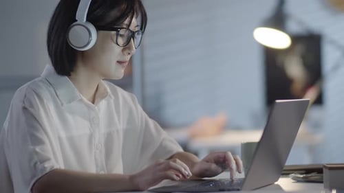 Asian Woman in Headphones Working in Office in Evening