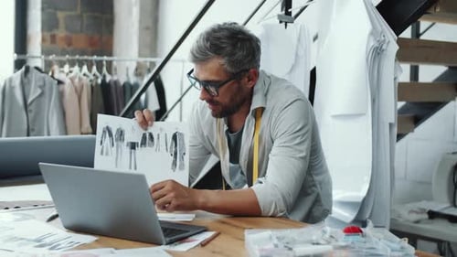 Male Designer Discussing Fashion Illustrations on Video Call in Sewing Studio