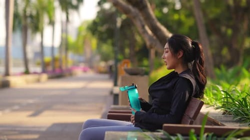 4K Asian woman drinking water from a bottle while jogging at public park in the morning.