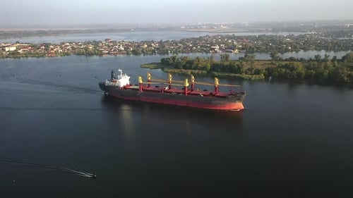 Luftaufnahme in Zeitlupe auf ein im Hafen ankommendes Frachtschiff, das an Bäumen und kleinen Gebäuden vorbeifährt