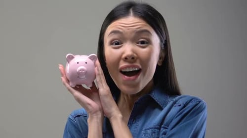 Lächelnde asiatische Frau schüttelt Sparschwein, Tipps zum Geldsparen, Finanzen und Budget