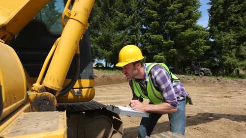 Construction Worker Inspecting Yellow Excavator Outdoors