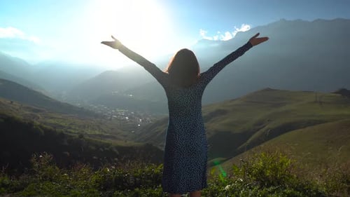 Eine junge Frau steht in einem Kleid und hebt ihre Hände hoch und blickt auf die Berge. Die Sonne scheint