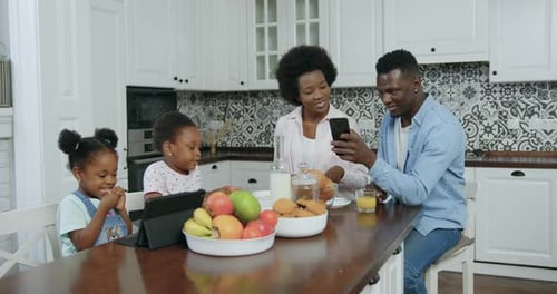 Pareja de piel negra revisando aplicaciones en el teléfono y sus dos hijas pequeñas que usan una tableta