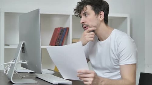 Young Adult Man Reviews Documents at Computer Desk