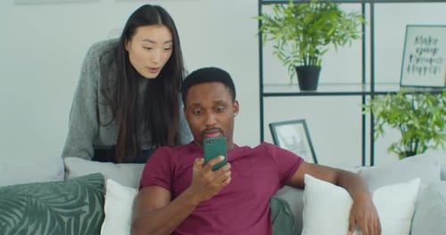 Happy Mixed Race Couple Talk Laugh Looking at Smartphone Using Funny Apps Sit on Couch