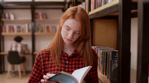 Young Redhead Teenage Girl in Red Shirt Standing Reading Book in Library and Smiling in Slowmotion