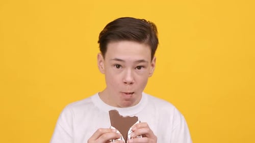 Happy Teen Boy Eating Chocolate Standing On Yellow Background