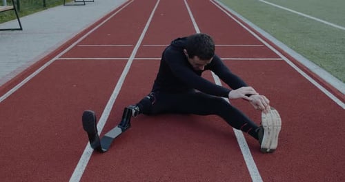 Disabled Man with Prosthetic Running Blades Stretching While Sitting at Sports Field