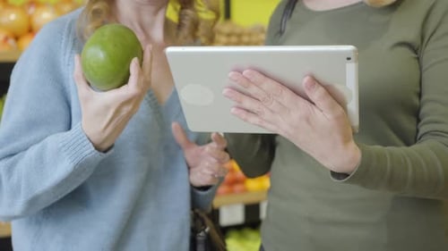 Two Unrecognizable Caucasian Women Using Tablet To Check Pomelo Origin in Grocery. Serious Ladies