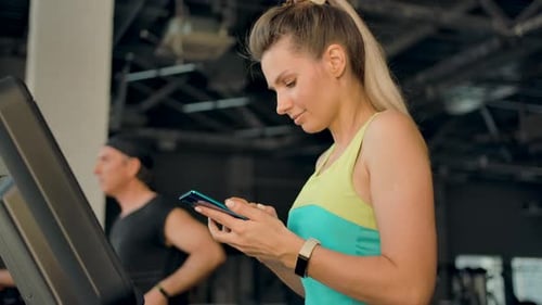 Blonde Woman Walking on a Treadmill in the Gym and is Typing a Message on Phone