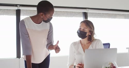 Two diverse female colleagues wearing face masks looking at laptop and discussing in office