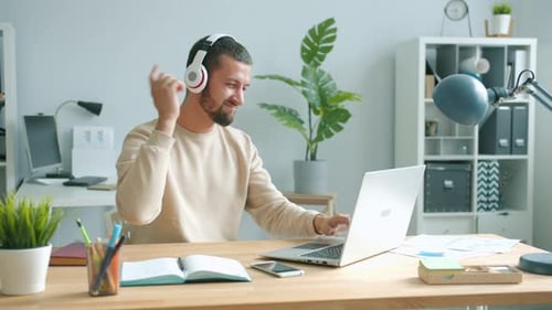 Cheerful Office Worker Wearing Headphones Moving Arms Dancing and Using Laptop