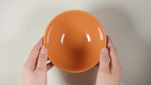 Menschliche Hände stellen eine orangefarbene Suppenschüssel auf einen weißen Tisch