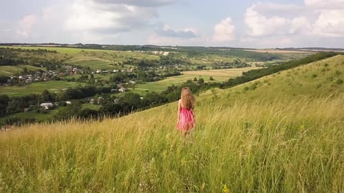Yong Woman with Long Hair in Red Dress Walking in Summer Field with Tall Green Grass on Rural Hill