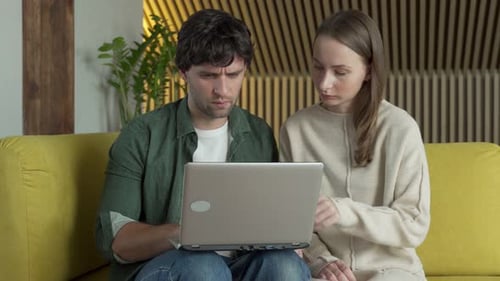 Junges Paar sitzt auf einem gemütlichen gelben Sofa und blickt auf einen Laptop-Bildschirm und gewinnt triumphierend