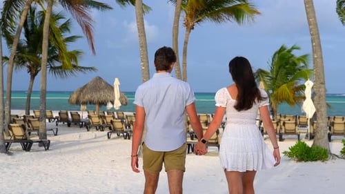 Flirtatious young couple walks on white sandy beach. Magic hour light with tropical palm trees and b