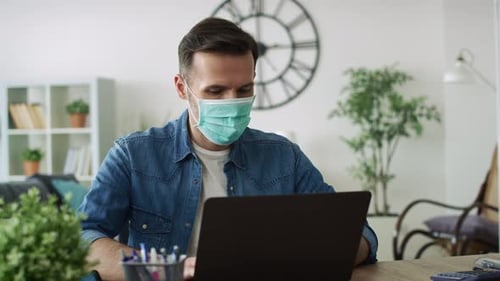 Man Works on Laptop at Home Wearing Mask