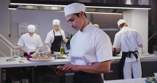 Mixed race male chef wearing chefs whites in a restaurant kitchen using a tablet and looking at came