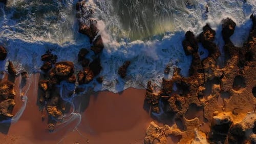 Aerial drone topdown shot of waves crashing the rocky beach shore, gazed by golden hour light