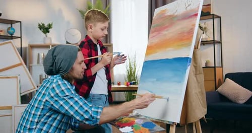 Artista masculino que enseña a un niño pequeño a dibujar en el estudio de su casa usando lienzo sobre caballete