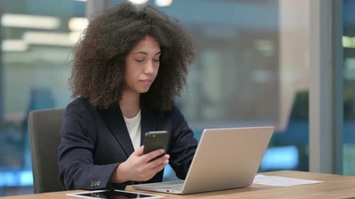 Woman Uses Smartphone at Desk with Laptop