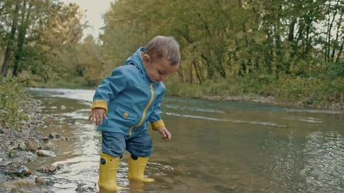 Cute Baby in Raincoat and Rubber Boots Has Fun By River, Throws Pebbles and Laughs. Funny Boy Learns