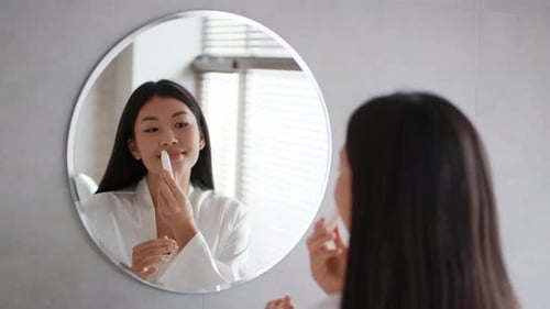 Asian Young Lady Applying Lipstick Looking At Mirror In Bathroom