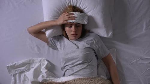 Femme bouleversée avec une compresse couchée dans son lit, souffrant de migraine prenant une pilule
