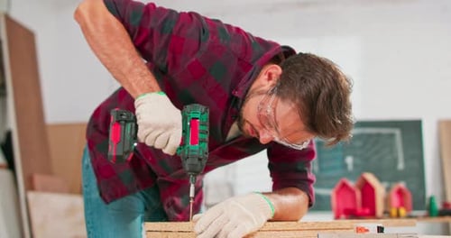 Concentrated Young Bearded Carpenter Wearing Jeans and a Plaid Flannel Shirt Drilling a Hole in a