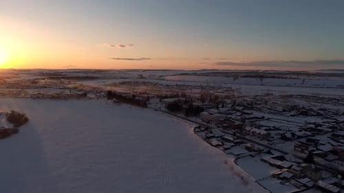 Aerial Over the Frozen Winding River Surrounded with Forest and with a Little Village on a Side