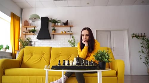 Young Woman Sitting on Yellow Sofa and Playing Chess in Room