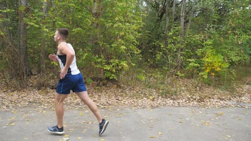 Young Strong Sportsman with Wireless Headphones Jogging Along Trail Near Forest