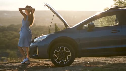 Young female driver standing near a broken car with open up hood inspecting her vehicle engine