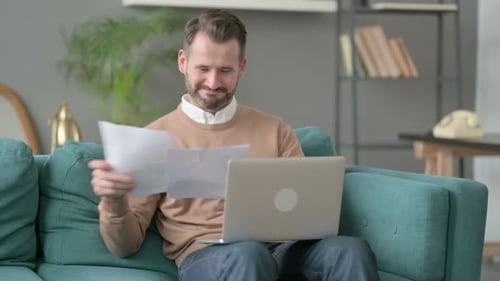 Man Sitting on Couch Reviews Paper With Laptop