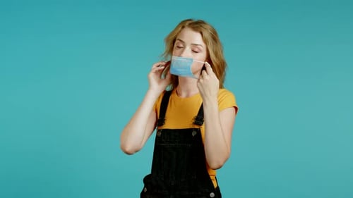 Young Pretty Teenager Girl Puts on Face Medical Mask During Coronavirus Pandemic. Portrait on Blue