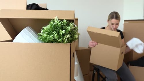 A Moving Woman Sits on a Chair and Puts Things Into a Cardboard Box in an Empty Apartment